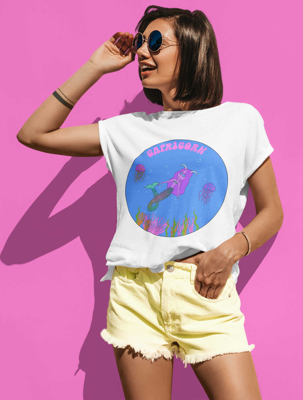 Cute girl with hippie glasses wearing a unisex capricorn t-shirt with a pink wall